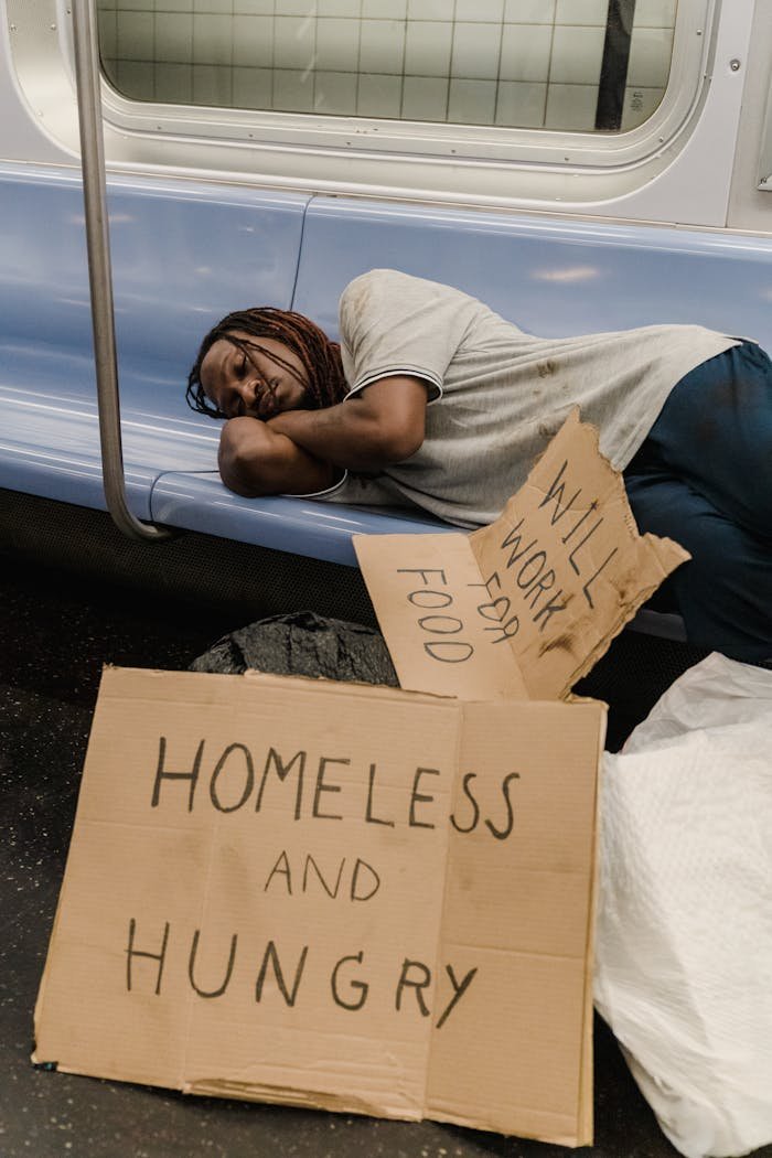 A man rests on a subway bench with cardboard signs reading Homeless and Hungry.