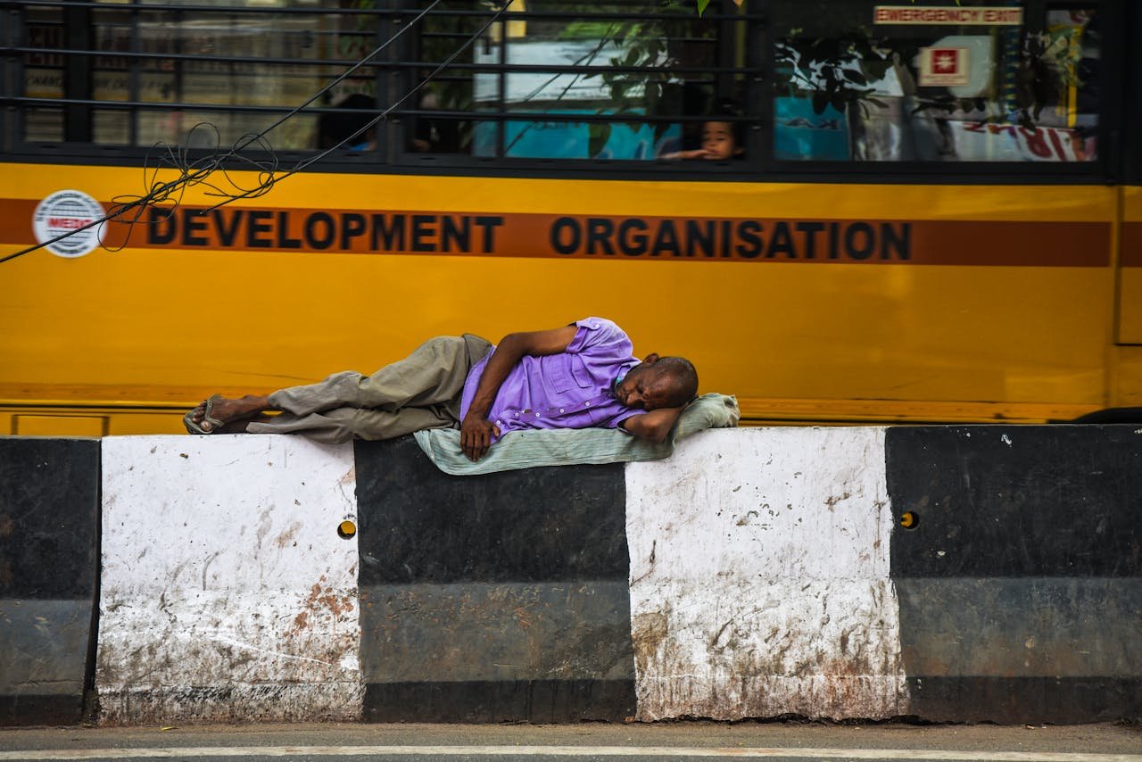 Elderly man sleeping on a concrete barricade beside a moving yellow bus in an urban setting.